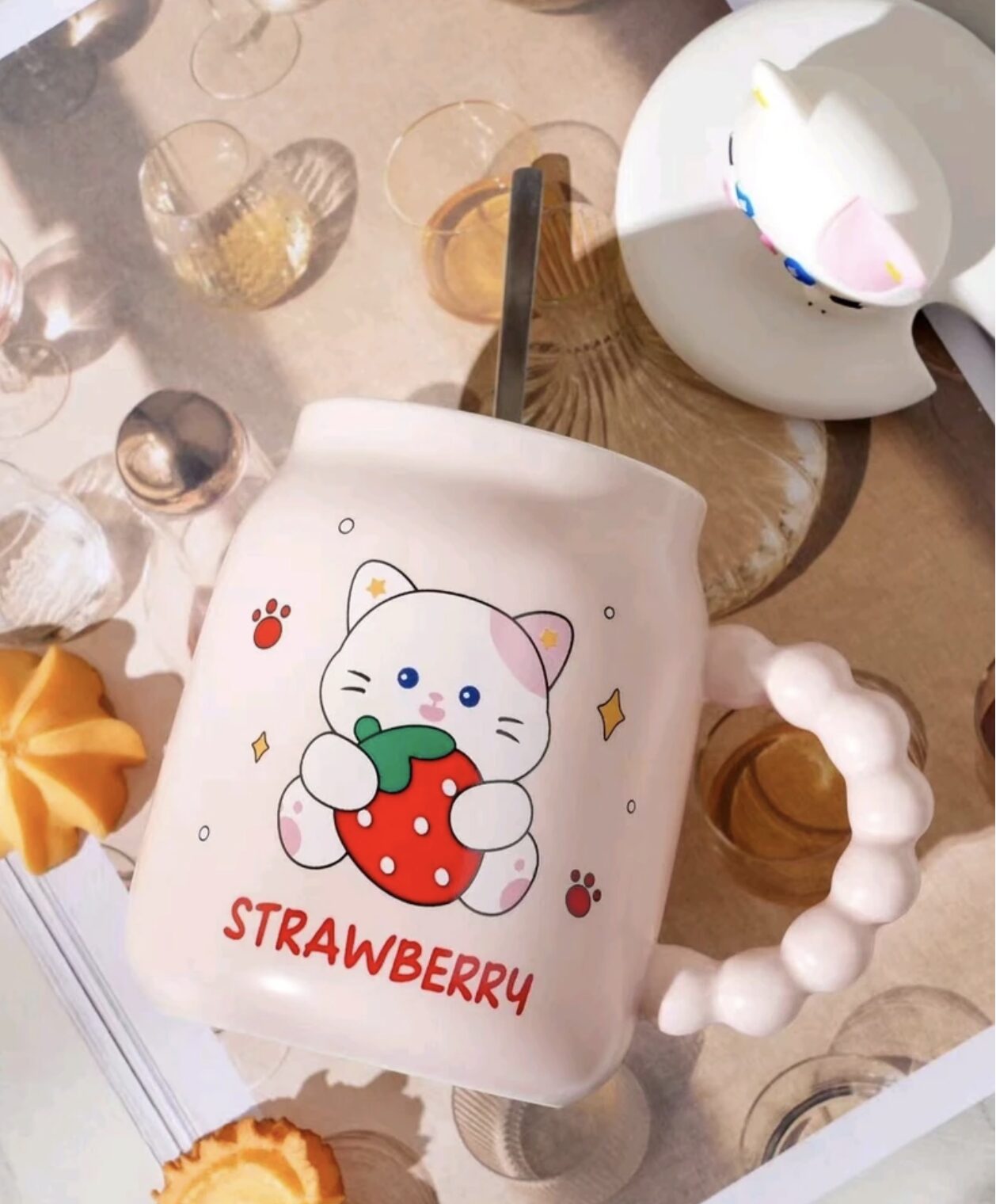 Taza mug color crema con diseño de gatito sosteniendo fresa, tapa 3D blanca y mango texturizado. Incluye cuchara y accesorios decorativos.